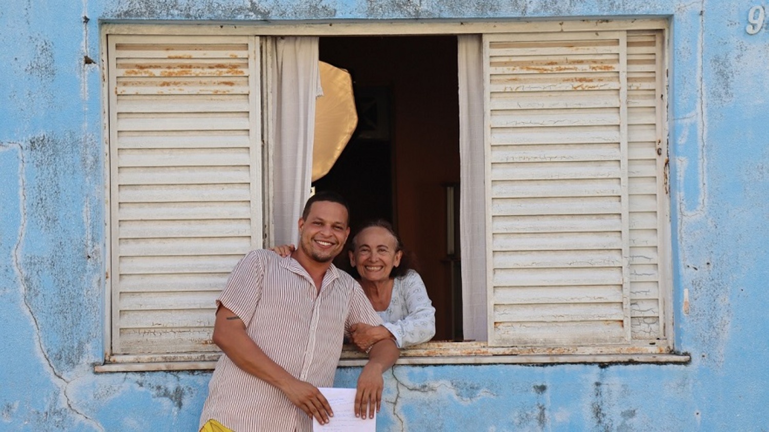 Imagem de divulgação de O Armário de Gisélia: os dois personagens abraçados junto a uma janela de uma casa azul
